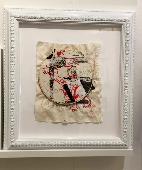 Mixed media artwork featuring a circular embroidery hoop on textured fabric with layered text and colorful scribbles. Framed in a white ornate frame.