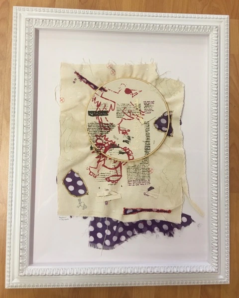 A framed mixed-media artwork featuring embroidered fabric with printed text, polka dot patterns, and an embroidery hoop.