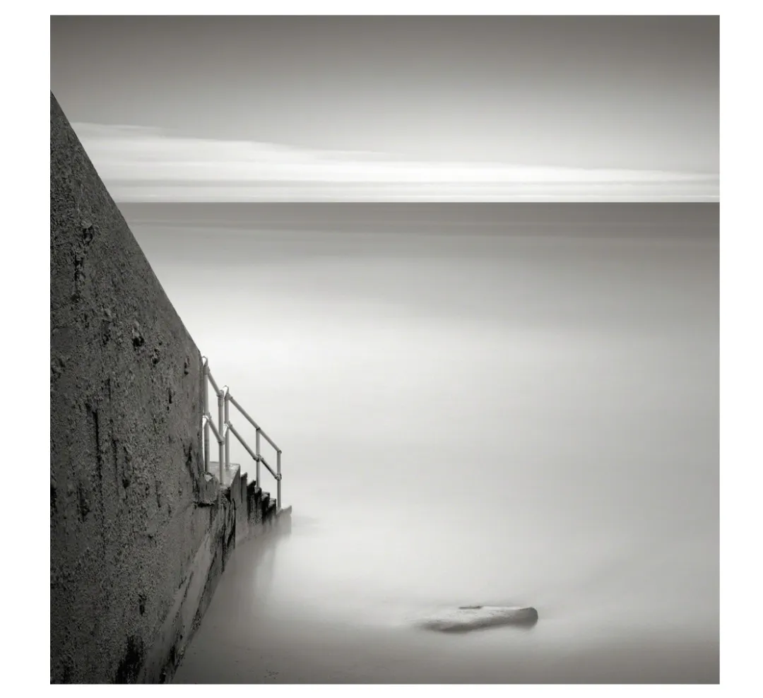 Black and white image of a stone stairway leading to a calm, misty sea under a cloudy sky, evoking tranquility and solitude.